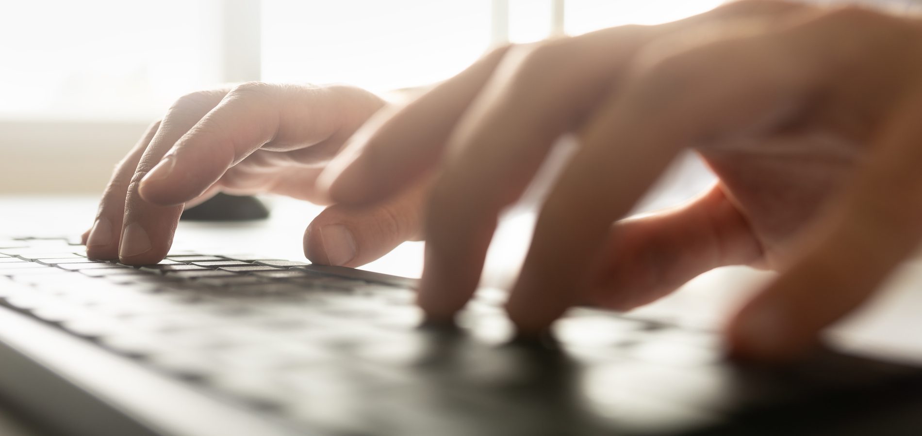 Low angle view of a businessman typing on a computer keyboard with a bright light flare coming in through a window over his hands.